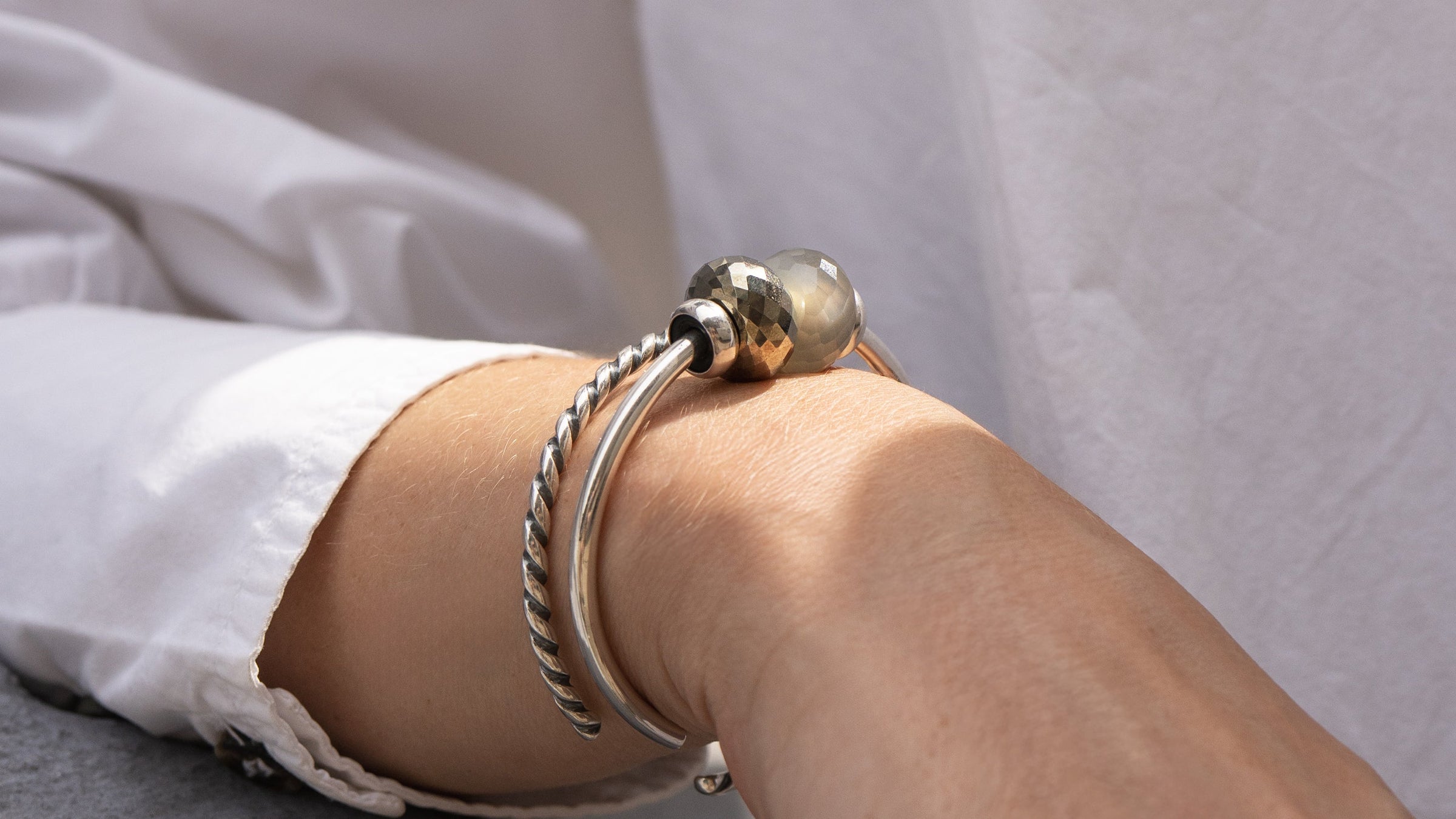 Close-up of a wrist wearing two silver bracelets with grey and golden beads on a white background